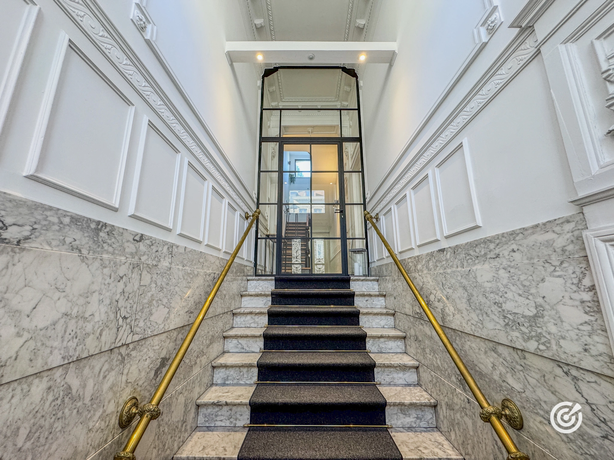 Élégant escalier en marbre recouvert d'un tapis sombre menant à une porte vitrée à verrière noire dans un grand hall d'entrée.