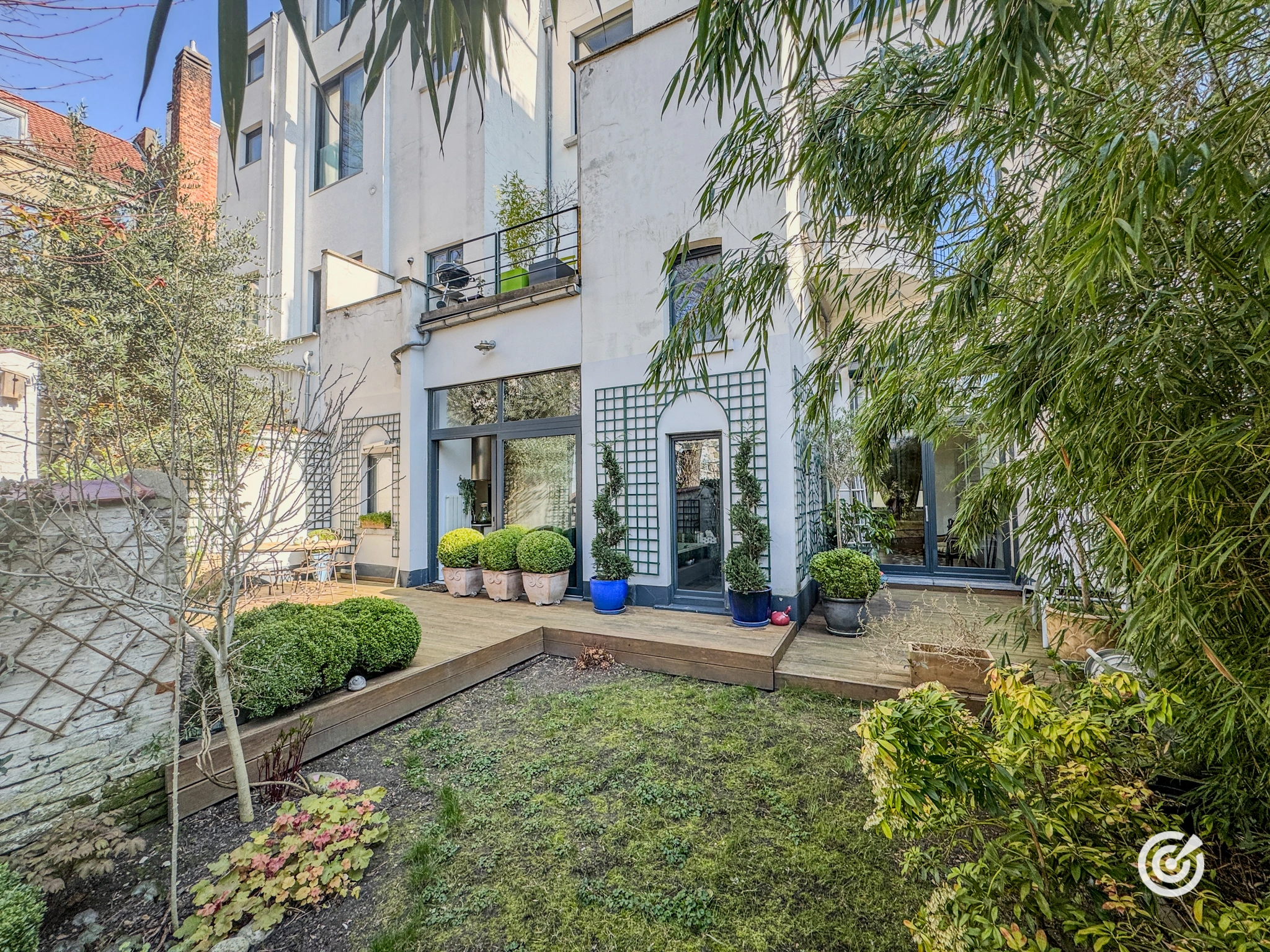 Jardin privatif paysager avec terrasse en bois, arbustes soigneusement entretenus et façade arrière d’une maison de ville moderne dotée de grandes baies vitrées et d’un balcon.